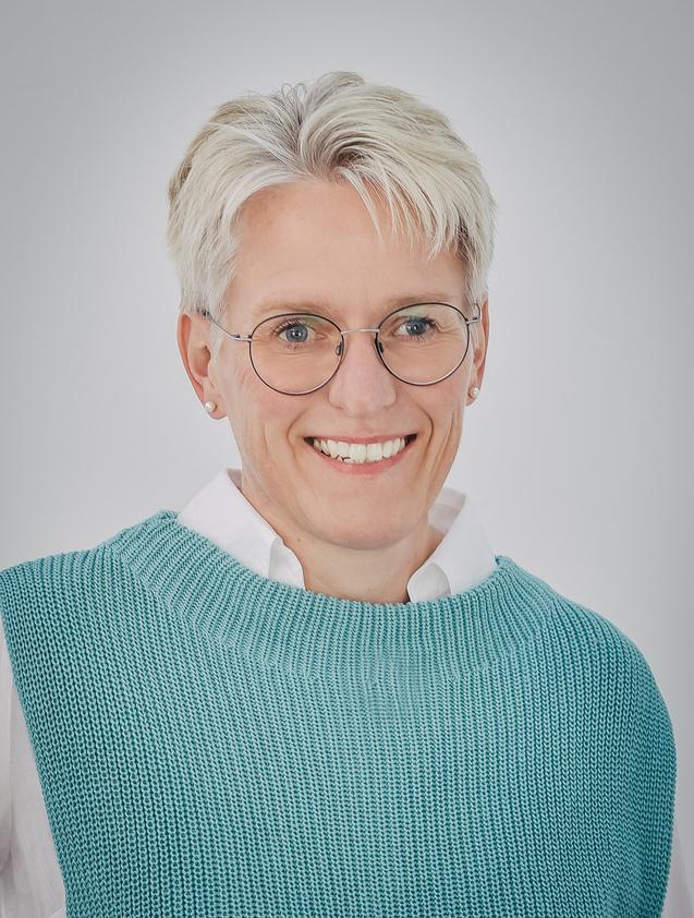 Eva Witthake Lächelnde Frau mit kurzen, blondierten Haaren, Brille und türkisfarbenem Pulli vor neutralem Hintergrund.